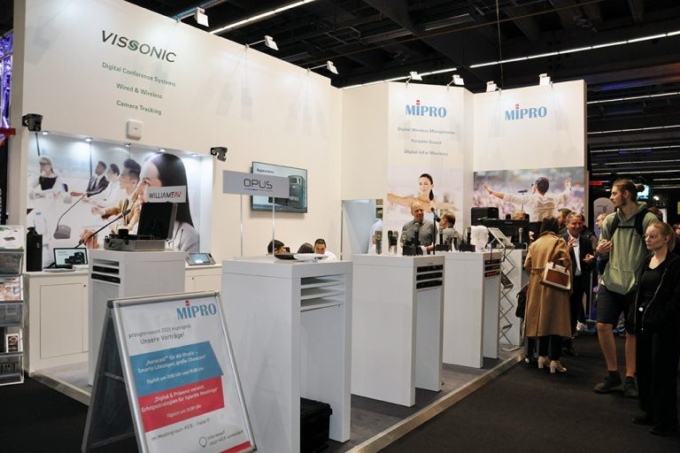 Der Messestand von MIPRO auf der Prolight + Sound 2025 präsentiert sich hell, offen und professionell. Im Zentrum stehen mehrere Produktinseln mit digitaler Drahtlostechnik – darunter Mikrofone, Taschensender und In-Ear-Systeme. Ein aufgestelltes Rollup kündigt spannende Vorträge zu digitalen Mikrofonsystemen, smarter Bühnenpraxis und erfolgreicher Medientechnik an. Die große Besucherzahl am Stand sowie das rege Interesse an den präsentierten Lösungen unterstreichen die Relevanz von MIPRO im professionellen Audiomarkt. Begleitet wird die Ausstellung von auffälligen Visuals, die die Einsatzbereiche der Technik – von Business bis Bühne – eindrucksvoll in Szene setzen.