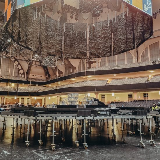 Das Bild zeigt den Aufbau einer Bühne in einer großen Veranstaltungshalle, der Festhalle Frankfurt. Zu sehen ist eine runde Bühnenkonstruktion, die auf zahlreichen höhenverstellbaren Gerüstfüßen mit Rollen montiert ist. Über der Bühne hängen schwere Vorhänge oder akustische Elemente sowie Teile der Licht- und Tontechnik. Im Hintergrund sind die Sitzreihen und die markante Architektur der Halle zu erkennen. Es handelt sich um einen technischen Aufbauprozess, bei dem die Bühne noch nicht vollständig fertiggestellt ist.