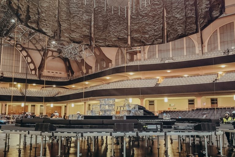 mott-mobile-systems-fitzek-festhalle Das Bild zeigt den Aufbau einer Bühne in einer großen Veranstaltungshalle, der Festhalle Frankfurt. Zu sehen ist eine runde Bühnenkonstruktion, die auf zahlreichen höhenverstellbaren Gerüstfüßen mit Rollen montiert ist. Über der Bühne hängen schwere Vorhänge oder akustische Elemente sowie Teile der Licht- und Tontechnik. Im Hintergrund sind die Sitzreihen und die markante Architektur der Halle zu erkennen. Es handelt sich um einen technischen Aufbauprozess, bei dem die Bühne noch nicht vollständig fertiggestellt ist.