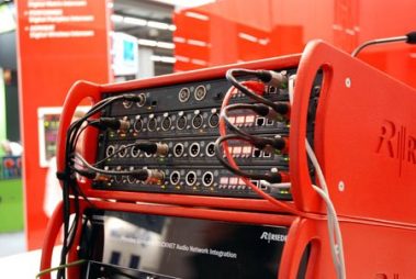 Das Bild zeigt eine technische Audiokonfiguration auf einer Messe. Im Vordergrund steht ein leuchtend rotes Rack mit zahlreichen Audiogeräten, das mit verschiedenen Kabeln verbunden ist. Diese Geräte, vermutlich Audio-Interfaces oder Signalprozessoren, verfügen über zahlreiche Eingangs- und Ausgangsanschlüsse. Die Kabel sind sorgfältig angeschlossen, was auf eine Demonstration oder Testeinrichtung hinweist. Im Hintergrund ist die Messeumgebung zu erkennen, die durch rote Wände und Informationsschilder geprägt ist. Das Logo des Herstellers „Riedel“ ist gut sichtbar und deutet auf professionelle Audiotechnologie hin. Die Szene vermittelt einen Eindruck von moderner Technik und zeigt ein Beispiel für Audio-Netzwerkintegration im professionellen Bereich.