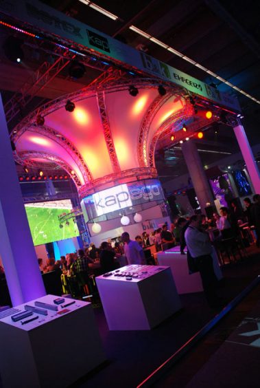 Das Bild zeigt einen beeindruckenden Messestand, der durch eine auffällige Beleuchtung und ein modernes Design hervorsticht. Die zentrale Struktur besteht aus einem geschwungenen, beleuchteten Dach, das in warmen Farben wie Rot und Orange erstrahlt und von einem stabilen Gerüst aus Metallstreben getragen wird. Im Hintergrund sind Besuchergruppen zu sehen, die sich an Stehtischen unterhalten. Auf einer großen Leinwand wird ein Fußballspiel gezeigt, was für eine dynamische und interaktive Atmosphäre sorgt. Im Vordergrund befinden sich Präsentationstische mit ausgestellten Produkten, die den Besuchern nähergebracht werden. Die Umgebung ist in ein farbenfrohes Licht gehüllt, das durch verschiedene Scheinwerfer erzeugt wird, was den Stand besonders lebendig und einladend wirken lässt.