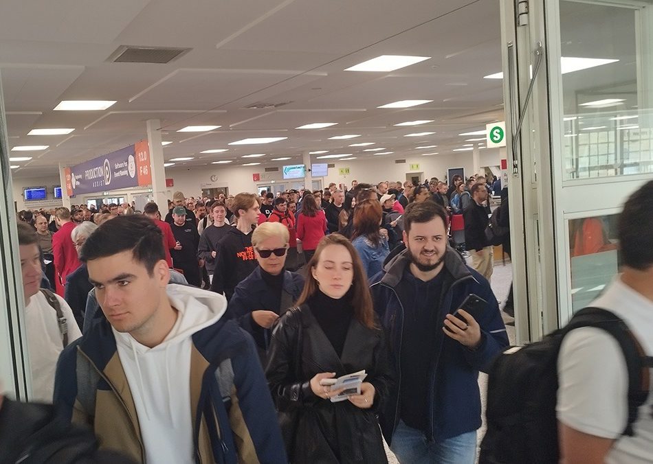 The picture shows the busy entrance to a trade fair or event hall, apparently just before or after the event has started. Hundreds of people can be seen streaming through the wide doors and into the hall; many of them are holding tickets, smartphones or brochures. The atmosphere appears busy yet orderly. In the background, there is a large banner bearing the words 'Production + DJ' and hall numbers (e.g. 12.0, F 46), which indicates that the event is a trade fair – presumably Prolight + Sound 2025. Signposts and monitors to help visitors find their way around, as well as 'S' signs (perhaps for 'service' or 'shuttle'), reinforce the impression of a major event that has been organised to a high standard. The image captures the moment of the rush, with many visitors, some in groups and some alone, making their way to the exhibition areas. It demonstrates the high level of interest in the event.
