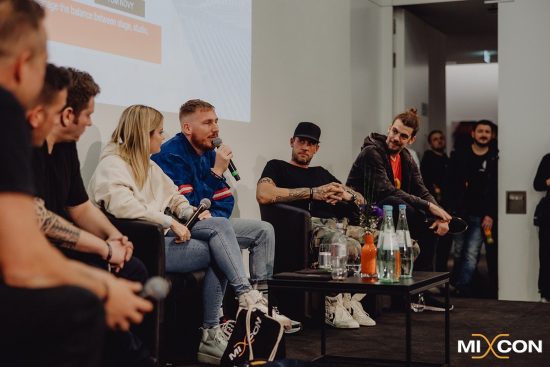 Das Bild zeigt eine Podiumsdiskussion auf der MIXCON x BVD DJ- & Producer Conference. Mehrere Personen sitzen auf einer Bühne in einem modernen Konferenzraum. Eine Person in einer blauen Jacke mit roten Streifen spricht in ein Mikrofon, während die anderen Panelisten aufmerksam zuhören. Die Atmosphäre wirkt entspannt und professionell. Im Vordergrund ist ein Tisch mit Wasserflaschen und Blumen zu sehen. Im Hintergrund stehen einige Zuschauer, die das Gespräch verfolgen. Das MIXCON-Logo in der unteren rechten Ecke des Bildes weist auf den Veranstaltungszusammenhang hin.