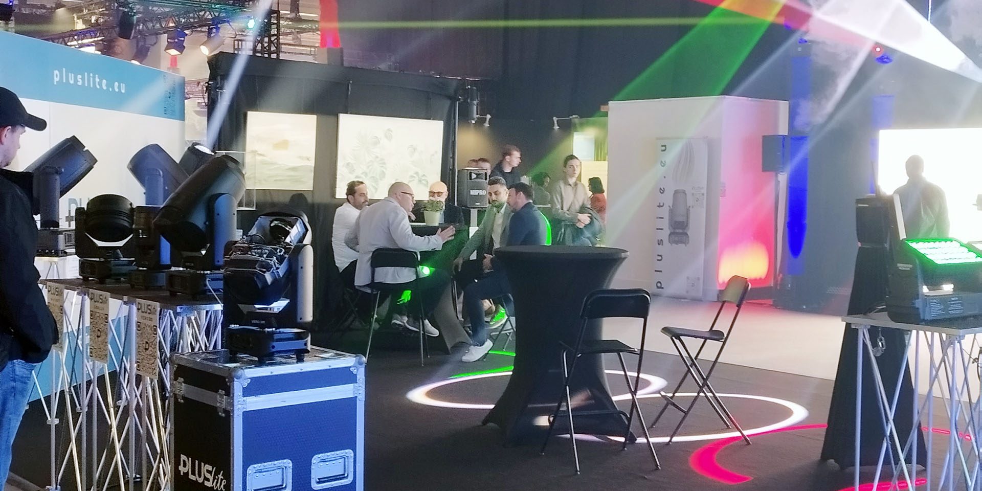 Messestand von Pluslite mit Scheinwerfern, Lichtstrahlen und Besuchergruppe in Gesprächen.