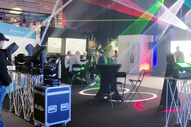 pluslite-pls-25 Messestand von Pluslite mit Scheinwerfern, Lichtstrahlen und Besuchergruppe in Gesprächen.