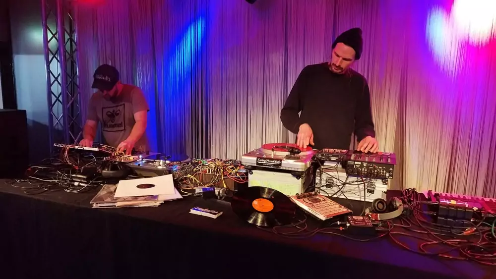 The image shows two DJs or music producers working at a large table with extensive equipment. The scene is bathed in atmospheric, red-blue stage lighting. The DJ on the left is wearing a cap labelled 