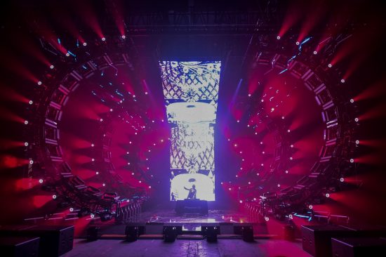 DJ in front of LED wall, surrounded by red moving lights and circular stage.