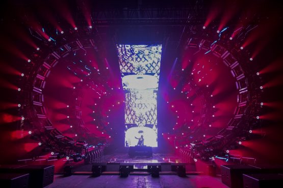 DJ in front of LED wall, surrounded by red moving lights and circular stage.