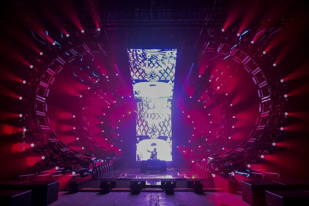 DJ in front of LED wall, surrounded by red moving lights and circular stage.