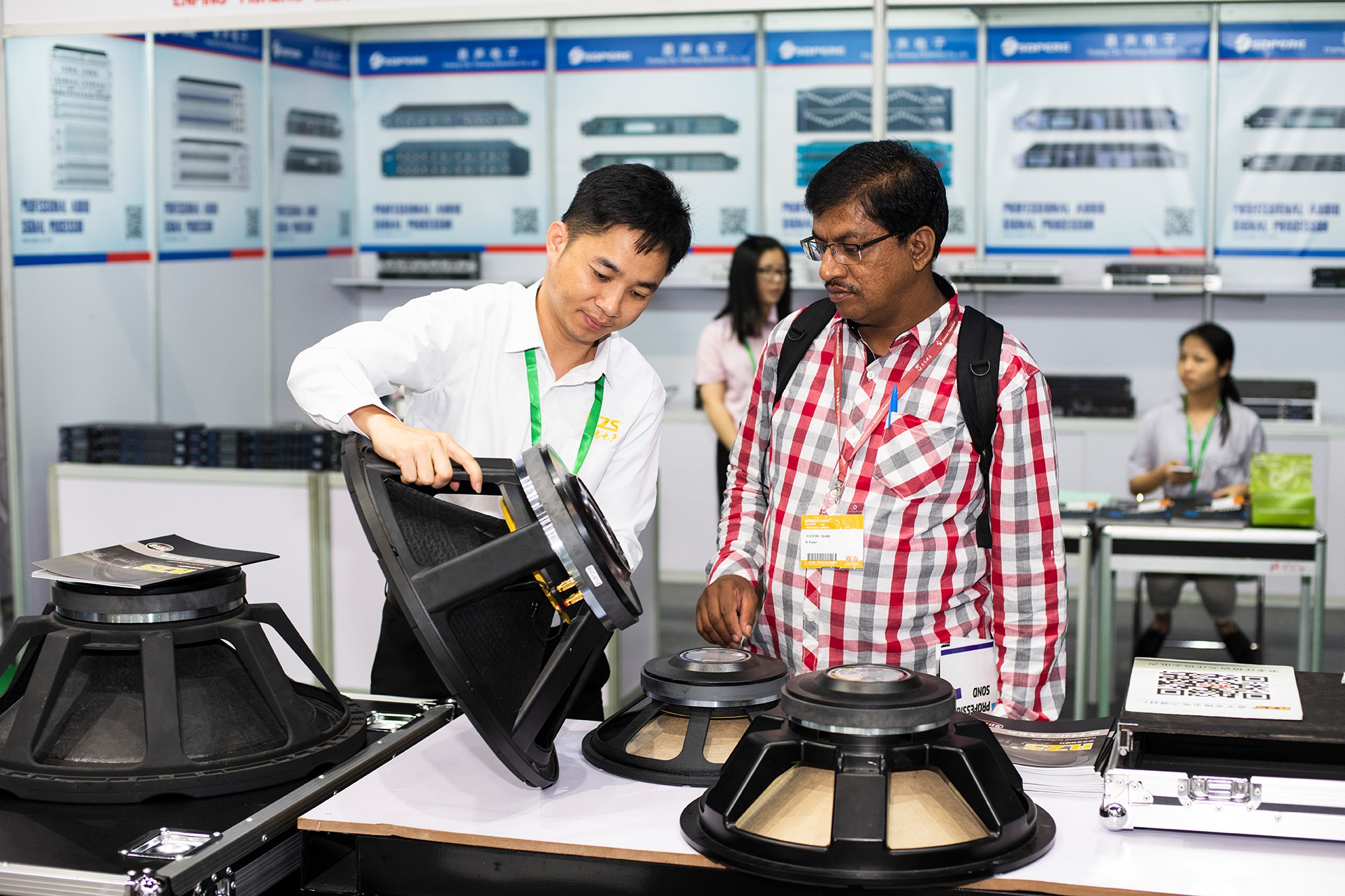 Two men look at large loudspeaker components at a trade fair stand for audio technology.