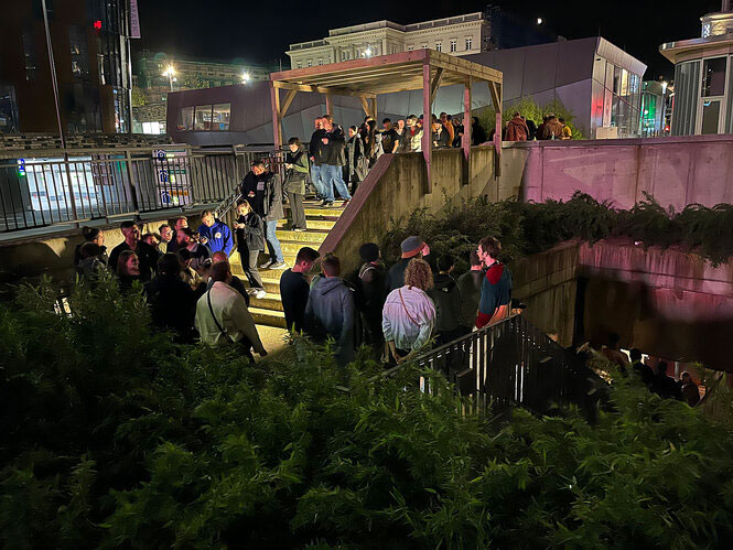 Das Bild zeigt eine größere Menschenmenge, die sich auf einer Treppe und in einem Eingangsbereich versammelt hat. Die Szenerie spielt sich nachts ab, und die Umgebung ist durch künstliches Licht beleuchtet. Die Personen wirken entspannt, einige unterhalten sich, während andere sich umsehen oder auf den Eingang zusteuern. Die Architektur deutet auf eine urbane Umgebung hin, mit Betonwänden, Geländern und einer überdachten Eingangsstruktur. Im Hintergrund sind moderne Gebäude und Lichter zu sehen, die eine lebendige Nachtatmosphäre schaffen. Möglicherweise handelt es sich um den Einlass zu einer Veranstaltung oder einem Club.