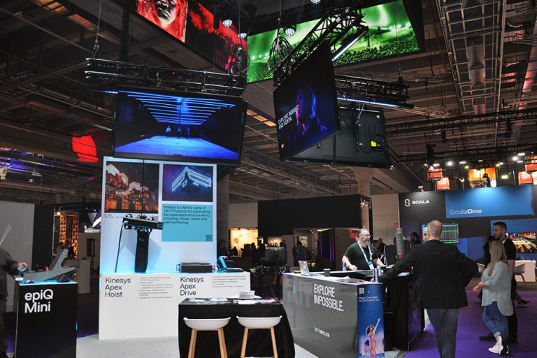tait-pls-25 Messestand von Tait mit epiQ Mini Console, Bildschirmen und Besucherinnen und Besuchern auf Messe.