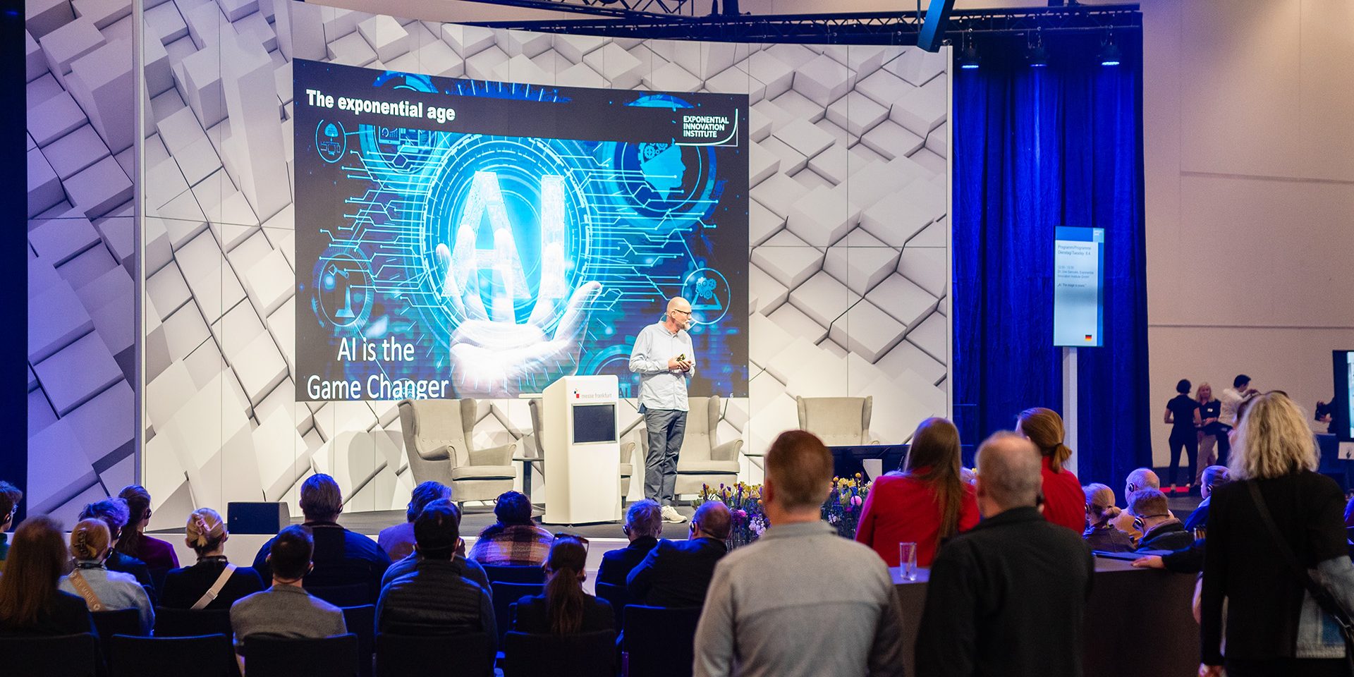 uve-samuels-ai-gamechanger Ein Redner präsentiert auf einer Bühne zum Thema KI vor einem aufmerksam lauschenden Publikum.
