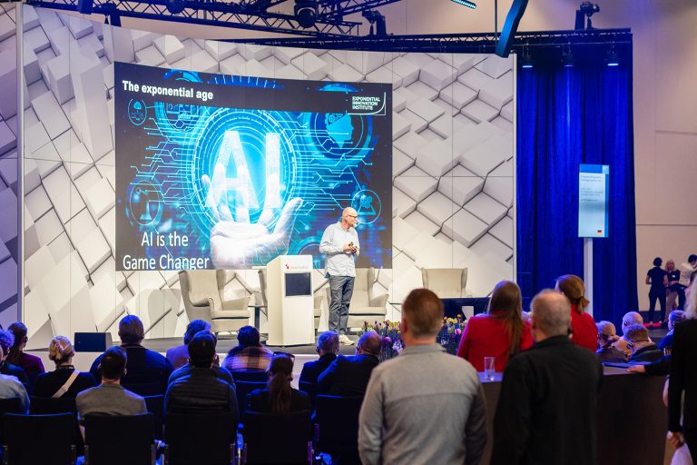 Ein Redner präsentiert auf einer Bühne zum Thema KI vor einem aufmerksam lauschenden Publikum.