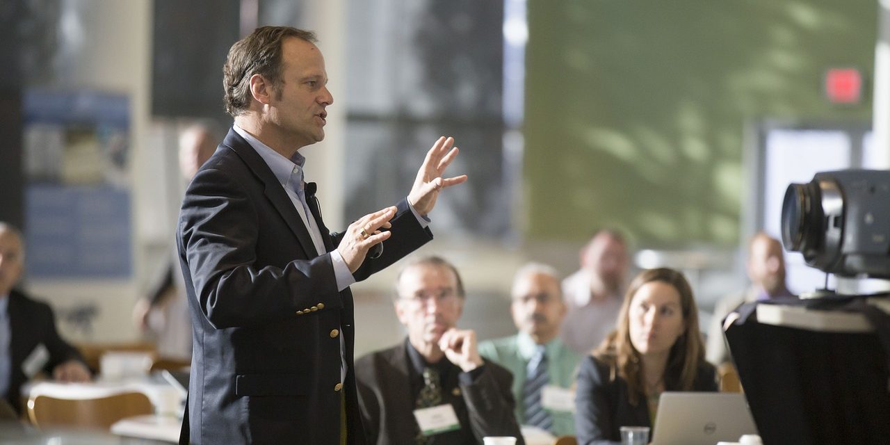 vote2
Das Bild zeigt eine Szene, in der ein Mann in einem dunklen Anzug eine Präsentation oder einen Vortrag hält. Er steht im Vordergrund und gestikuliert mit den Händen, während er spricht. An seinem Revers ist ein Mikrofon befestigt, was auf eine professionelle Veranstaltung hindeutet. Im Hintergrund sitzen mehrere Personen an runden Tischen und hören aufmerksam zu. Einige Teilnehmer haben Notebooks oder Unterlagen vor sich, was darauf hinweist, dass es sich um ein Meeting, eine Konferenz oder ein Seminar handelt. Auf der rechten Seite ist eine Kamera zu sehen, die möglicherweise den Vortrag aufzeichnet. Die Atmosphäre wirkt geschäftlich und professionell.