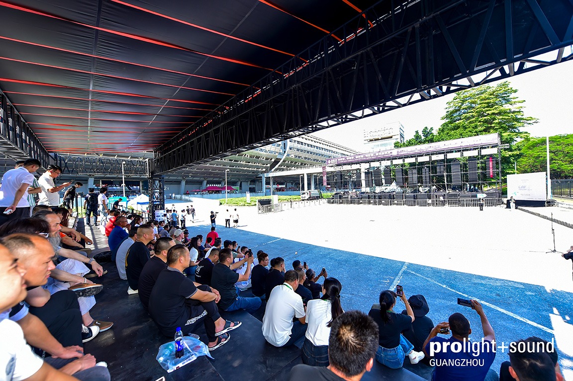 Spectators watch an outdoor demonstration featuring a large stage and line array systems